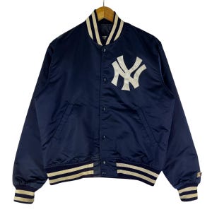 Può includere: Una giacca da baseball in satin blu navy con un logo ricamato bianco dei New York Yankees sul retro. La giacca ha un colletto, polsini e orlo a costine.