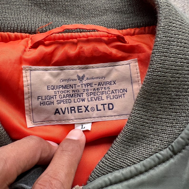 May include: An olive green flight jacket with a bright orange lining and a ribbed collar. A rectangular label is sewn inside, featuring the text Certificate Authenticity, EQUIPMENT-TYPE-AVIREX, STOCK NO. 28-A6755, FLIGHT GARMENT SPECIFICATION, HIGH SPEED LOW LEVEL FLIGHT, AVIREX LTD, and a size L tag.