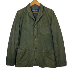 May include: Olive green blazer with three buttons, two front pockets, and a chest pocket. The blazer has a notched lapel and a label that reads "COMME des GAR&Ccedil;ONS HOMME". The jacket is hanging on a wooden hanger.