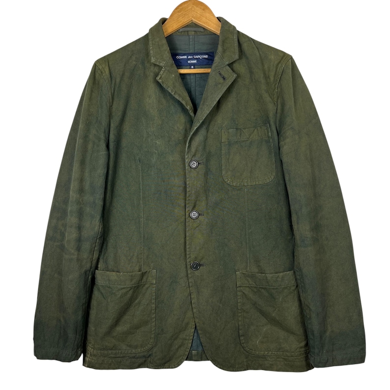 May include: Olive green blazer with three buttons, two front pockets, and a chest pocket. The blazer has a notched lapel and a label that reads "COMME des GAR&Ccedil;ONS HOMME". The jacket is hanging on a wooden hanger.