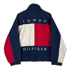 May include: Navy blue and red Tommy Hilfiger jacket with white accents. The back of the jacket features the brand name "TOMMY" above a white, red, and white color block, and "HILFIGER" below. The jacket has a zipper closure and a stand-up collar.