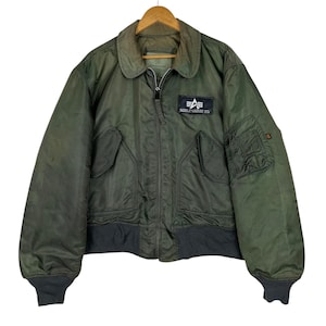May include: Olive green bomber jacket with a black ribbed hem and cuffs. Features a zip closure, two front pockets, and a patch on the chest. The jacket is hanging on a wooden hanger.