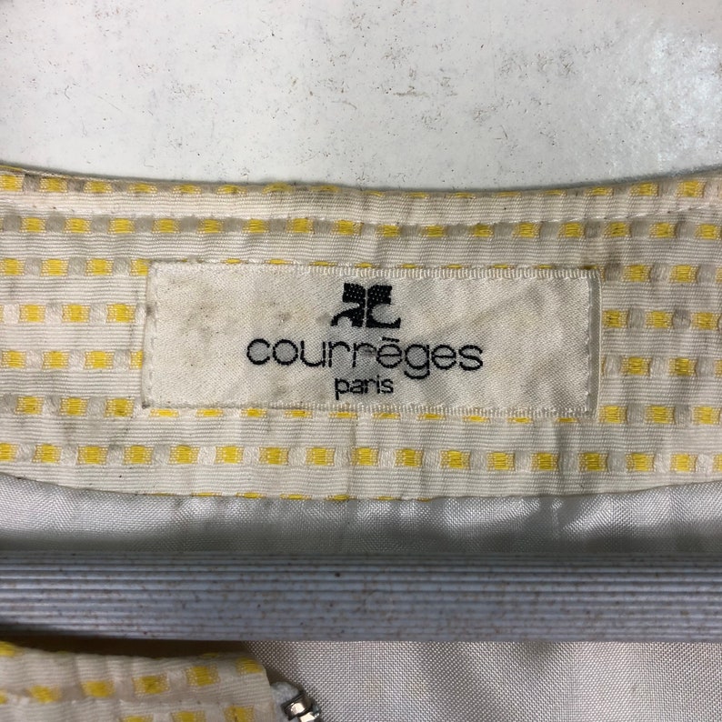 K&ouml;nnte beinhalten: Ein wei&szlig;es Etikett mit schwarzem Text, der "Courr&egrave;ges Paris" lautet, mit einem stilisierten "C"-Logo &uuml;ber dem Text. Das Etikett ist an einem wei&szlig;en Stoff mit gelbem Karomuster befestigt.