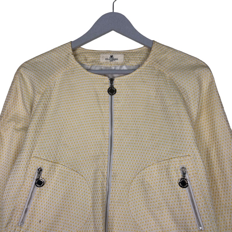 K&ouml;nnte beinhalten: Eine gelbe Jacke mit wei&szlig;em Muster, Rei&szlig;verschluss und Logo auf der Vorderseite. Die Jacke hat zwei Taschen mit Rei&szlig;verschl&uuml;ssen auf der Vorderseite.