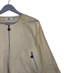 K&ouml;nnte beinhalten: Eine wei&szlig;e Jacke mit gelbem Muster, Rei&szlig;verschluss und kleinem Logo auf der Tasche. Die Jacke hat einen runden Ausschnitt und lange &Auml;rmel.