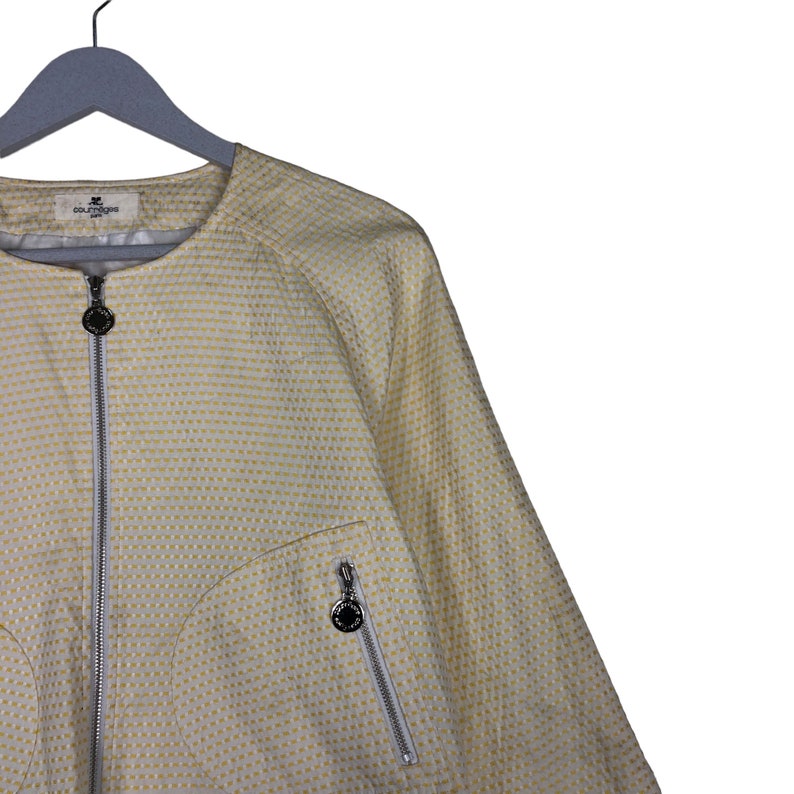 K&ouml;nnte beinhalten: Eine wei&szlig;e Jacke mit gelbem Muster, Rei&szlig;verschluss und kleinem Logo auf der Tasche. Die Jacke hat einen runden Ausschnitt und lange &Auml;rmel.