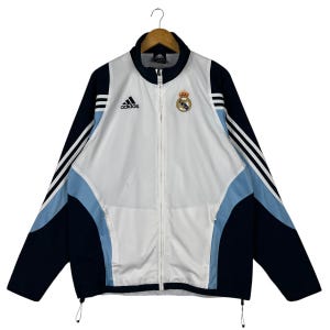 May include: A white, light blue, and navy blue Adidas track jacket with a full zipper. The jacket features the Adidas logo and the Real Madrid crest on the chest. The sleeves have white stripes.