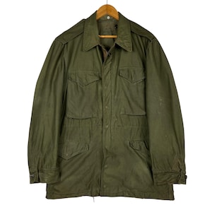 May include: An olive green military-style jacket with four front pockets and a collared neckline. The jacket is suspended on a wooden hanger. The jacket features a button-up front and long sleeves.