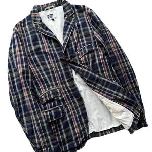 Puede incluir: Una blazer a cuadros en azul marino, rojo y verde oliva. La chaqueta tiene solapas con muescas, dos bolsillos delanteros y cierre de dos botones. El forro interior es blanco. La blazer está hecha de tela ligera.