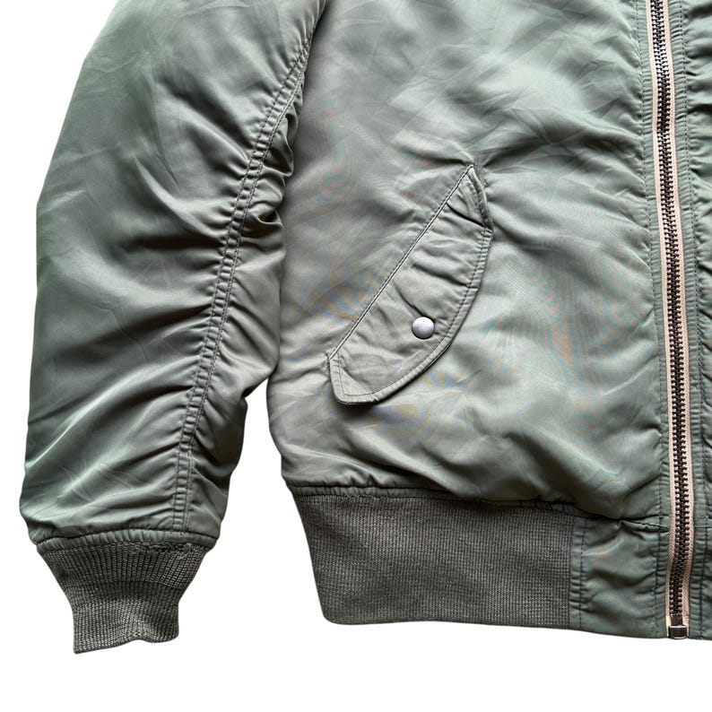 May include: An olive green bomber jacket featuring a smooth nylon-like exterior, a front metal zip closure, a single slanted side pocket with a snap button flap, and ribbed knit cuffs and hem for a secure fit around the waist and wrists.