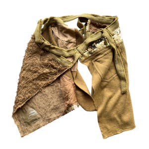 May include: A unique, deconstructed garment featuring a mix of textures and colors. The piece combines olive green fabric, a fuzzy brown material, and sections adorned with sequins. It has a wrap-around design with tie closures.