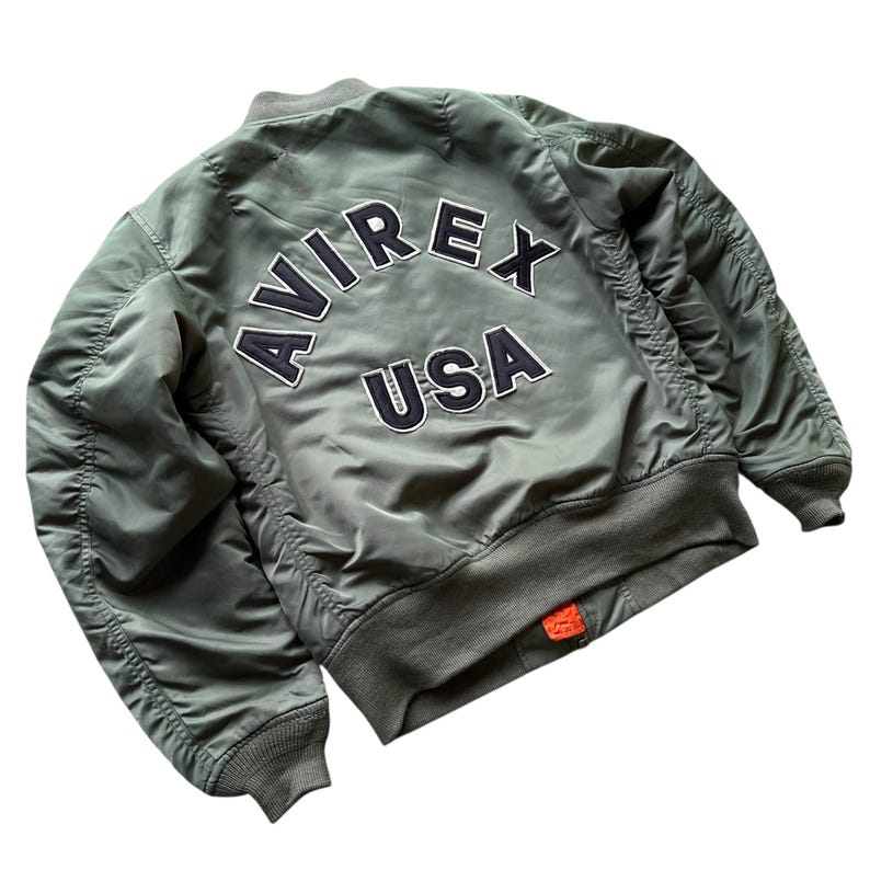 May include: An olive green bomber jacket featuring a ribbed collar, cuffs, and hem. The back displays the text AVIREX USA in large, dark navy blue embroidered letters with white trim. The jacket has long sleeves and a smooth, nylon-like finish.
