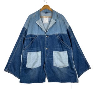 Könnte beinhalten: Eine Jeansjacke im Patchwork-Design. Die Jacke hat einen hellblauen oberen Bereich und Ärmel, einen dunkelblauen unteren Bereich und zwei hellblaue quadratische Taschen. Sie hängt an einem Holzbügel.