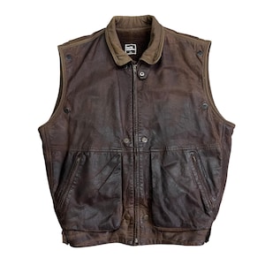 May include: A brown leather vest with a zip-up front, two front pockets, and a collar. The vest has a vintage look with visible wear and tear. The vest has a label that reads "Isaac Mizrahi".