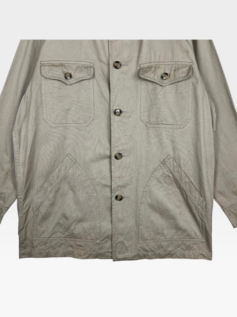 K&ouml;nnte beinhalten: Beige Jacke mit vier Vordertaschen und Kn&ouml;pfen. Die Jacke hat zwei Brusttaschen mit Knopfverschluss und zwei untere aufgesetzte Taschen. Die Kn&ouml;pfe sind dunkelbraun.