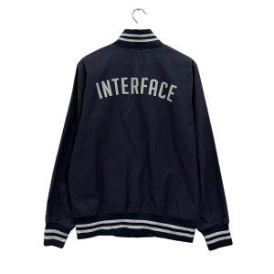 May include: Navy blue bomber jacket with the word "INTERFACE" in white lettering across the back. The jacket has a ribbed collar, cuffs, and hem with white stripes. The jacket is hanging from a metal hook.