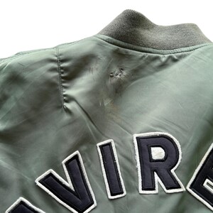 May include: A sage green bomber jacket featuring a ribbed collar and large black and white embroidered lettering across the back. The fabric shows a visible snag and slight discolouration near the shoulder seam, indicating wear on the garment.