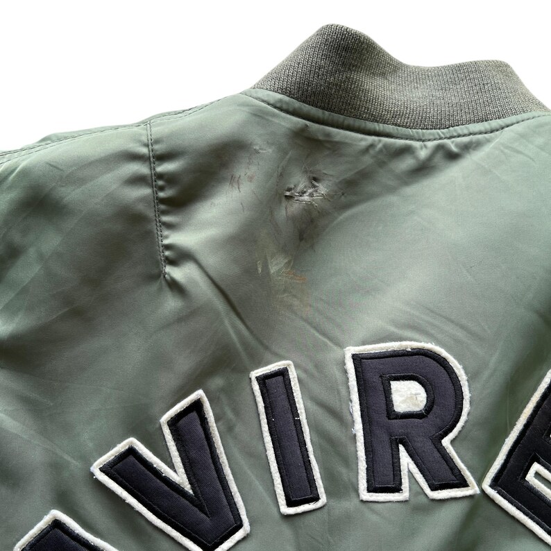 May include: A sage green bomber jacket featuring a ribbed collar and large black and white embroidered lettering across the back. The fabric shows a visible snag and slight discolouration near the shoulder seam, indicating wear on the garment.
