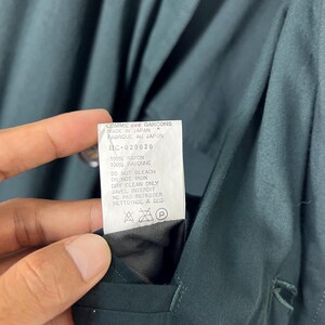 Może przedstawiać: Zbliżenie ciemnozielonego ubrania z guzikami i etykietą. Etykieta głosi "COMME des GARCONS MADE IN JAPAN" i zawiera instrukcje pielęgnacji. Tkanina to rayon.