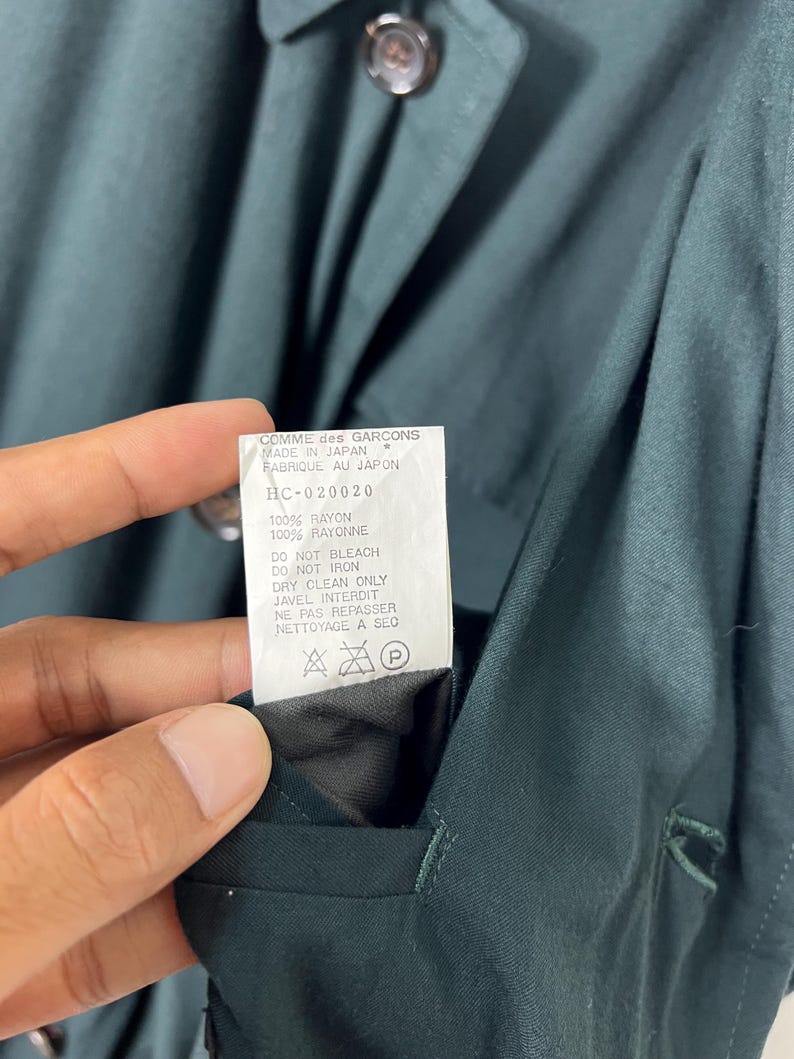 Może przedstawiać: Zbliżenie ciemnozielonego ubrania z guzikami i etykietą. Etykieta głosi "COMME des GARCONS MADE IN JAPAN" i zawiera instrukcje pielęgnacji. Tkanina to rayon.