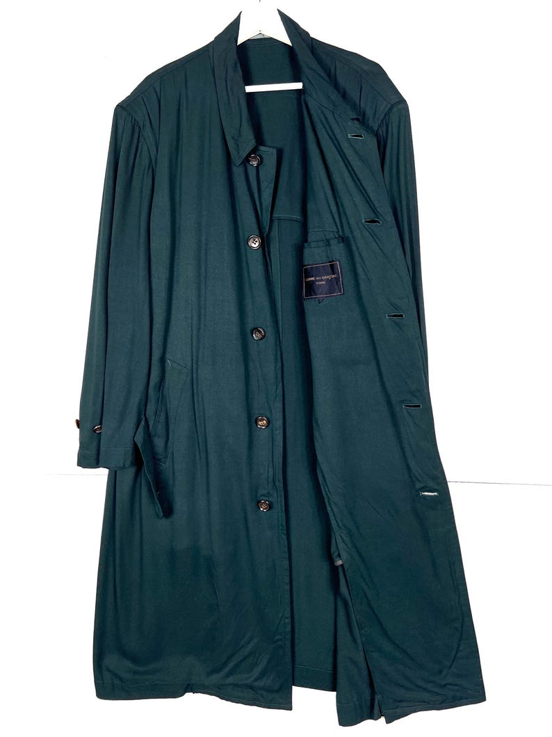 Może przedstawiać: Długi, ciemnozielony płaszcz z zapięciem na guziki i wykładanym kołnierzem. Płaszcz wisi na białym wieszaku na białym tle. Płaszcz ma kieszeń z metką w środku.