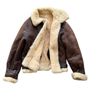 May include: A brown leather jacket with a cream shearling lining and collar. The jacket has a zip closure, a buckled waist, and shearling trim on the cuffs. The jacket is designed for warmth and protection from the elements, a classic outerwear piece.