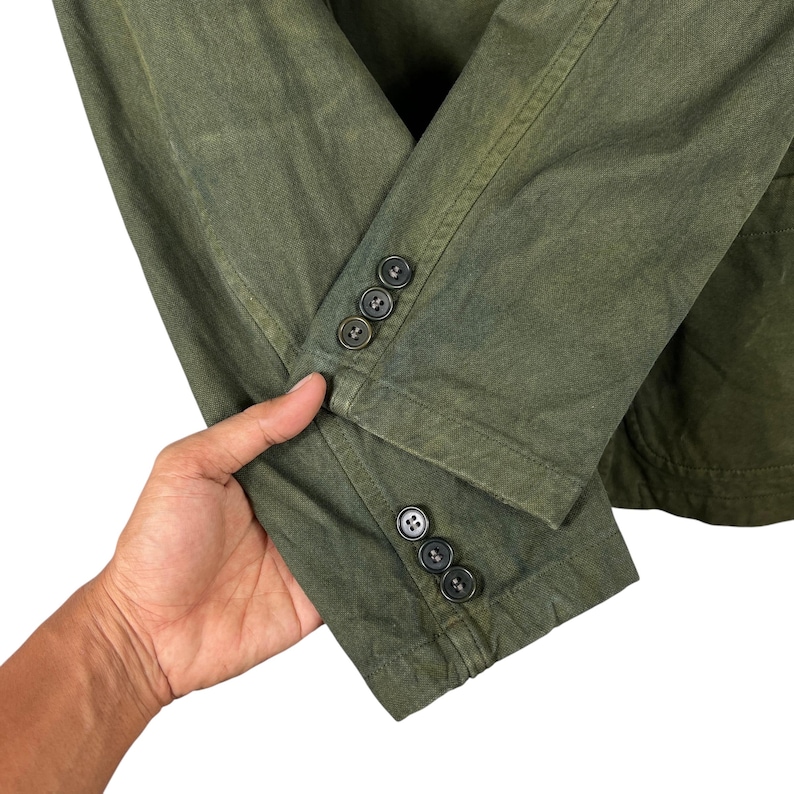 May include: Close-up of a green jacket sleeve with four dark buttons. The jacket has a textured appearance and a classic design. The sleeve is slightly wrinkled, suggesting a worn or vintage style. The buttons are evenly spaced.