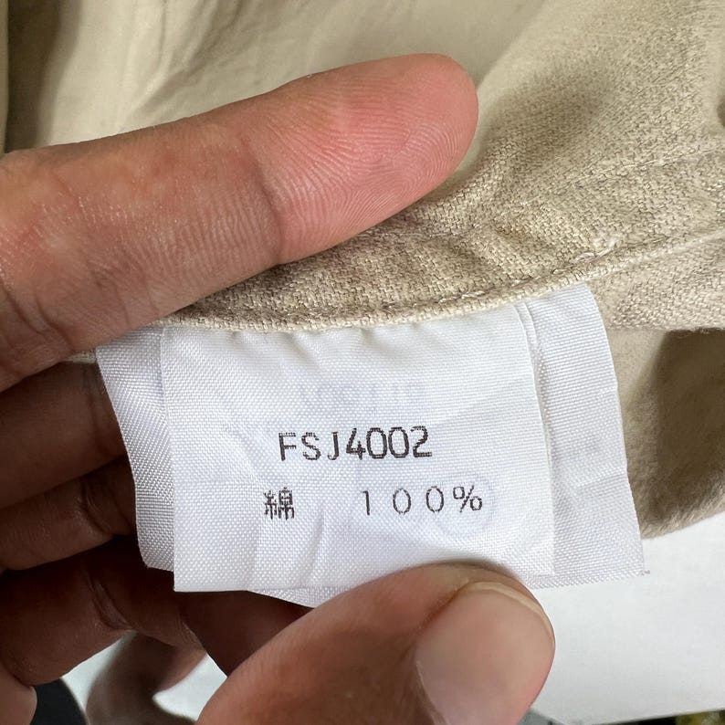 K&ouml;nnte beinhalten: Nahaufnahme eines Kleidungsetiketts mit dem Text "FSJ4002", "綿" und "100%". Das Etikett ist wei&szlig; mit schwarzem Text und an einem beigen Stoff befestigt. Der Stoff wird von einer Hand gehalten.