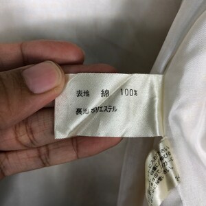 K&ouml;nnte beinhalten: Ein wei&szlig;es Stofflabel mit schwarzem Text, der "表地 綿 100% 裏地 ポリエステル" lautet, was mit "Obermaterial: Baumwolle 100% Futter: Polyester" &uuml;bersetzt wird.