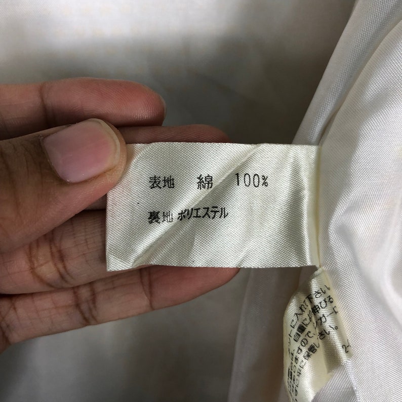 K&ouml;nnte beinhalten: Ein wei&szlig;es Stofflabel mit schwarzem Text, der "表地 綿 100% 裏地 ポリエステル" lautet, was mit "Obermaterial: Baumwolle 100% Futter: Polyester" &uuml;bersetzt wird.