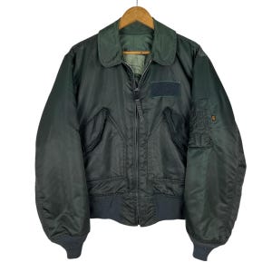 May include: A dark green bomber jacket with a zip-up front, two front pockets, and a patch on the chest. The jacket has a ribbed collar and cuffs. The jacket is hanging on a wooden hanger.