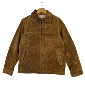 Könnte beinhalten: Braune Wildlederjacke mit durchgehendem Reißverschluss und zwei Brusttaschen. Die Jacke hat einen Kragen und ein weiches, hellfarbiges Futter. Sie hängt an einem Holzbügel.