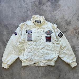Könnte beinhalten: Eine cremefarbene Honda F1-Rennjacke mit hohem Kragen, langen Ärmeln mit gerippten Bündchen und einem Frontreißverschluss. Die Jacke ist mit verschiedenen gestickten Aufnähern verziert, darunter Honda-Schriftzüge, Grand-Prix-Logos und Rennteam-Embleme.