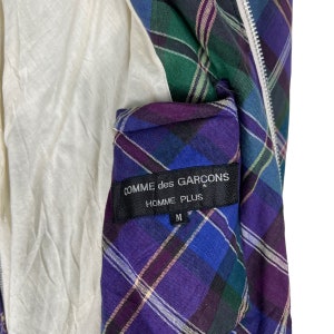 Puede incluir: Una chaqueta de cuadros de color morado, azul, verde y blanco con cierre de cremallera. La chaqueta tiene una etiqueta negra con el texto "COMME des GARCONS HOMME PLUS" y la talla "M" impresa en ella.