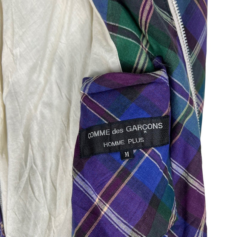 Puede incluir: Una chaqueta de cuadros de color morado, azul, verde y blanco con cierre de cremallera. La chaqueta tiene una etiqueta negra con el texto "COMME des GARCONS HOMME PLUS" y la talla "M" impresa en ella.