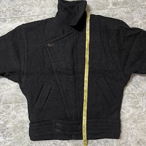 Puede incluir: Chaqueta de lana gris oscuro con cuello alto, cierre asim&eacute;trico con botones y cintura con cintur&oacute;n. La chaqueta tiene dos bolsillos delanteros y mide unos 69 cm de largo.