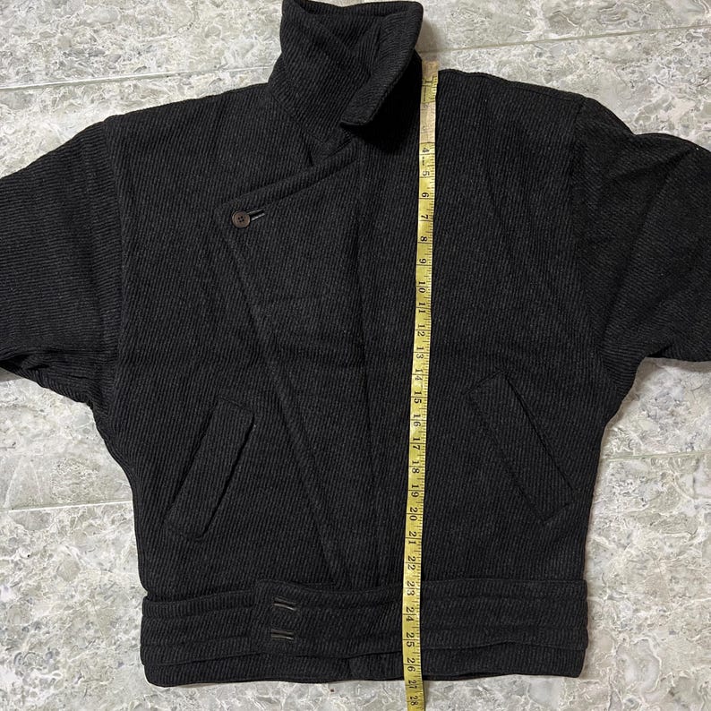 Puede incluir: Chaqueta de lana gris oscuro con cuello alto, cierre asim&eacute;trico con botones y cintura con cintur&oacute;n. La chaqueta tiene dos bolsillos delanteros y mide unos 69 cm de largo.