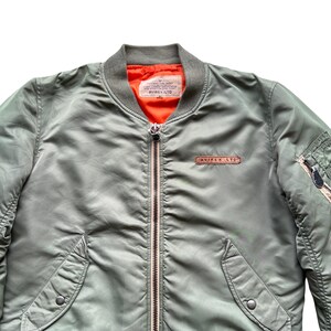 May include: An olive green bomber jacket featuring a bright orange interior lining, a ribbed collar, and a central metal zip. A rectangular leather patch with the text AVIREX LTD is on the chest, and a utility pocket with a zip is on the sleeve.