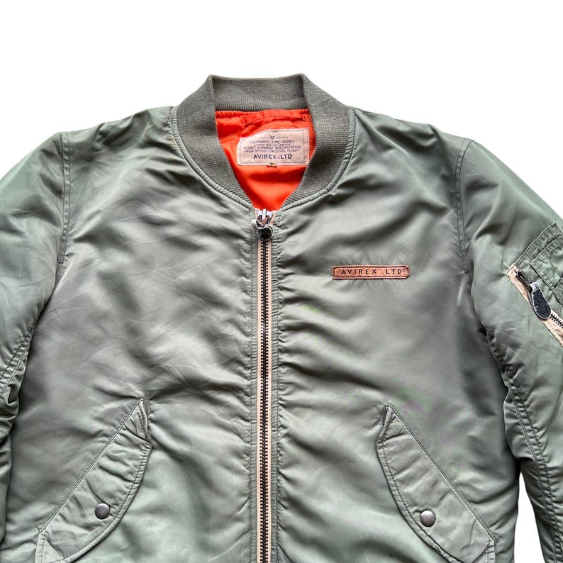 May include: An olive green bomber jacket featuring a bright orange interior lining, a ribbed collar, and a central metal zip. A rectangular leather patch with the text AVIREX LTD is on the chest, and a utility pocket with a zip is on the sleeve.