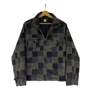 May include: A zip-up jacket featuring a patchwork design of square panels in shades of dark green, charcoal gray, and black. The jacket includes a high collar, two chest pockets with buttoned flaps, long sleeves with buttoned cuffs, and a dark plaid lining.