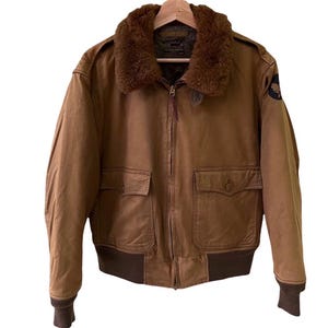 May include: A brown bomber jacket with a faux fur collar, two front pockets, and a zipper closure. The jacket has a ribbed hem and cuffs. A patch is visible on the left sleeve. The jacket is hanging on a wooden hanger.
