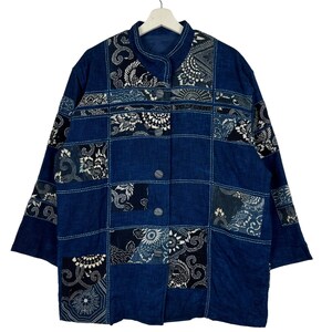 May include: A blue patchwork jacket with a mandarin collar and three-quarter sleeves. The jacket has a button-up front and is made of fabric squares with floral and geometric patterns in white and black. It hangs on a white hanger.