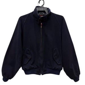 Könnte beinhalten: Marineblaue Jacke mit durchgehendem Reißverschluss, zwei Vordertaschen und Stehkragen. Die Jacke hängt an einem schwarzen Kleiderbügel vor weißem Hintergrund. Die Jacke hat einen gerippten Saum und Bündchen.