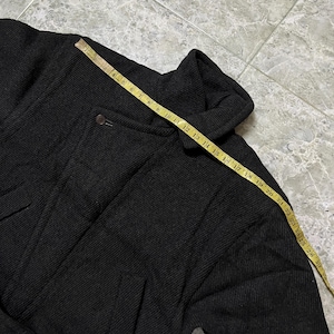 Puede incluir: Chaqueta gris oscuro con cuello alto y cierre de bot&oacute;n. Una cinta m&eacute;trica amarilla est&aacute; colocada sobre la chaqueta, mostrando medidas en cent&iacute;metros. La chaqueta tiene una tela texturizada.