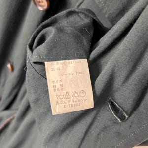 Może przedstawiać: Zbliżenie na ciemnozielony element odzieży z etykietą. Etykieta zawiera japoński tekst i instrukcje prania. Ubranie ma guzik i dziurkę na guzik. Tkanina wydaje się być rayonem.