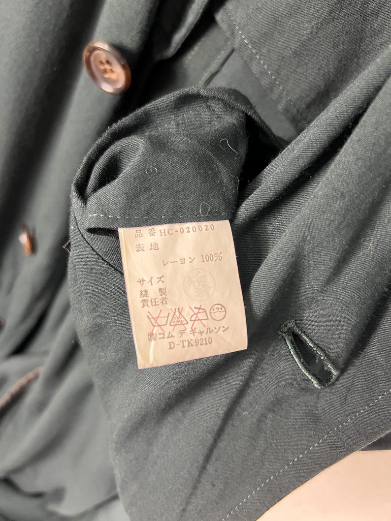 Może przedstawiać: Zbliżenie na ciemnozielony element odzieży z etykietą. Etykieta zawiera japoński tekst i instrukcje prania. Ubranie ma guzik i dziurkę na guzik. Tkanina wydaje się być rayonem.