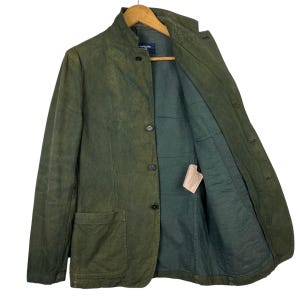 May include: Olive green blazer with a mandarin collar and button closure. The jacket has two front pockets and a dark green interior lining. The garment is hanging on a wooden hanger.