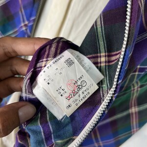 Puede incluir: Una etiqueta blanca con texto negro unida a una tela de cuadros p&uacute;rpura, verde y azul. La etiqueta dice "PJ-040880", "Polyester 100%", "100%", "100%", "Talla", "AD1997", "(株) コムデギャルソン", "D-TK9210".