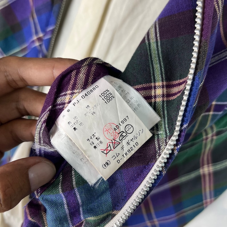 Puede incluir: Una etiqueta blanca con texto negro unida a una tela de cuadros p&uacute;rpura, verde y azul. La etiqueta dice "PJ-040880", "Polyester 100%", "100%", "100%", "Talla", "AD1997", "(株) コムデギャルソン", "D-TK9210".