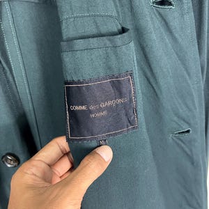 Może przedstawiać: Zbliżenie na ciemnozieloną kurtkę z kieszenią i etykietą "COMME des GARCONS HOMME". Kurtka ma zapięcie na guziki i metkę z rozmiarem "M". Tkanina wydaje się być gładkim, tkanym materiałem.